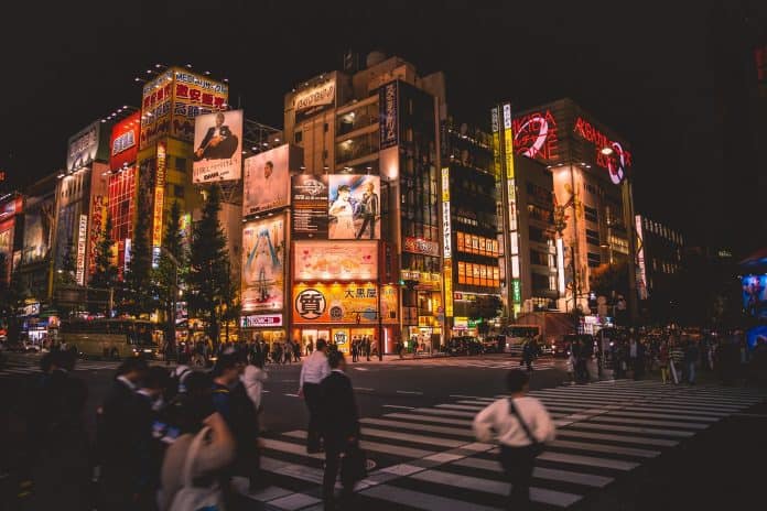 fotografia da cidade de Tóquio durante a noite. É possível visualizar prédios com diferentes outdoors no fundo da imagem, sendo que a frente pessoas cruzam uma faixa de pedestres.
