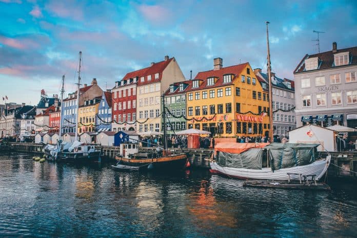 Fotografia da cidade de Copenhage, na qual é possível ver três barcos parados próximos a casas coloridas.