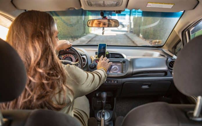 Fotografia de motorista mulher durante trabalho em aplicativo de transporte. Ela está acessando o celular, o banco do carona está vazio e o veículo está em uma rua com muros com vegetação dos dois, com movimentação de carros na via em frente