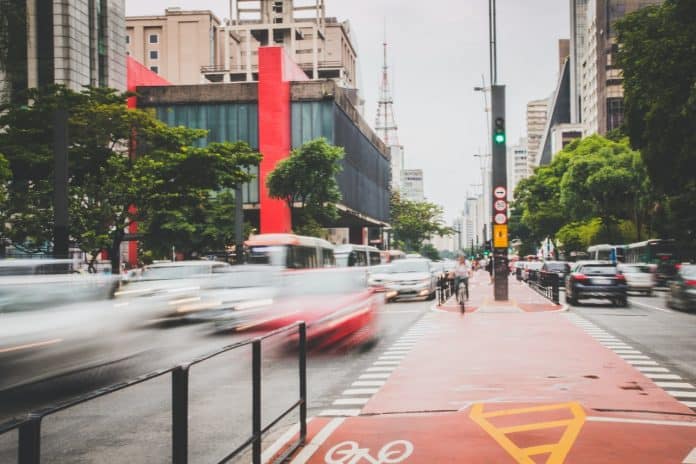 Fotografia da Avenida Paulista, em São Paulo, na altura do Masp, com circulação de veículos, pessoas andando de bicicleta na ciclovia, bem como edifícios e árvores