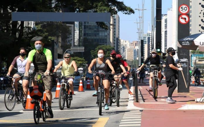 Foatografia de usuários, homens e mulheres, andando de bicicleta em ciclovia de avenida movimentada, em meio aos edifícios e também um pouco paisagismo, com algumas árvores