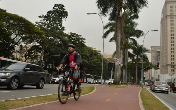 Fotografia de ciclovia na Avenida Brigadeiro Faria Lima, região oeste de São Paulo, com um usuário de bicicleta, onde se destacam a paisagem verde, edifícios e a movimentação de veículos