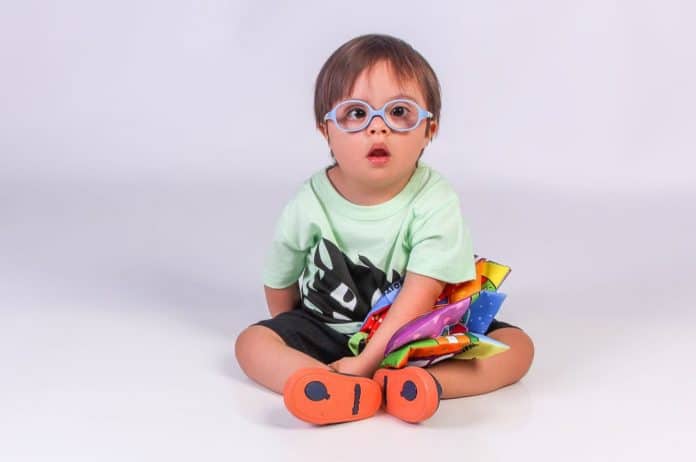 Criança durante ensaio fotográfico do projeto Galera do Click, realizado em 2020, para uma exposição sobre a síndrome de Down. Luis Fernando, 2 anos, na foto, sentado com um livro infantil colorido e roupa também colorida, nas cores verde e preto, tênis azul e laranja, e óculos azul