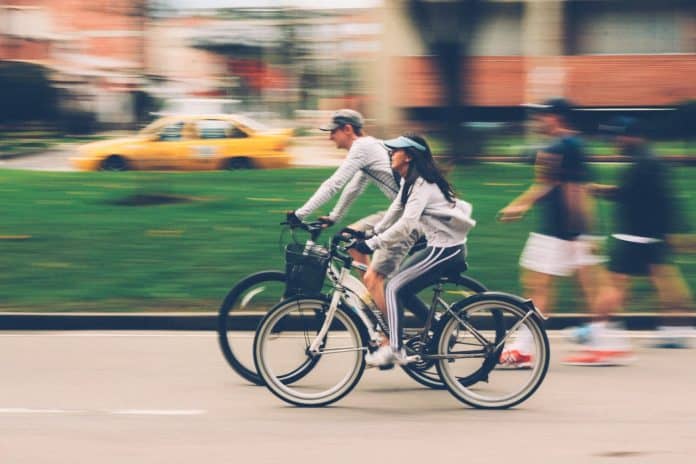 Fotografia de pessoas se locomovendo em centro urbano de bicicleta, caminhando e táxi, com área verde e, ao fundo, edificações