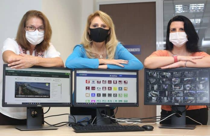 Fotografia de três mulheres da gestão do Departamento de Tecnologia da Informação da Prefeitura de Santos, São Paulo. Elas estão atrás de uma mesa com três computadores, que mostram imagens de monitoramento do município, e com os braços cruzados em cima dos mesmos